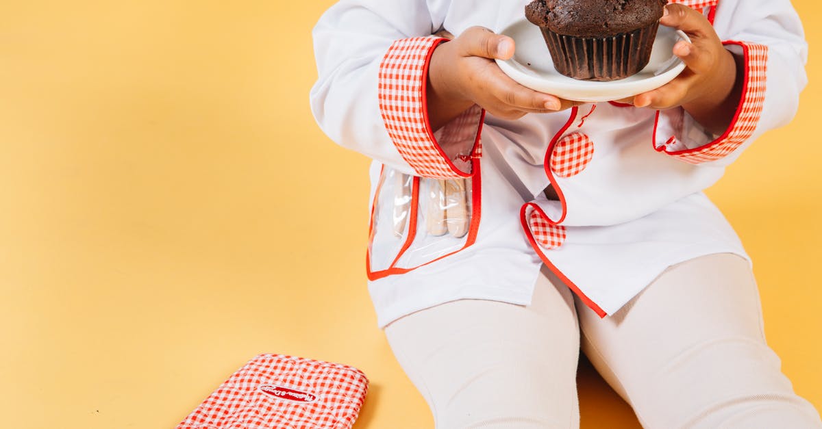 How can I bake muffins without an oven? - Crop anonymous ethnic little girl wearing cook uniform sitting with baked cupcake near oven glove against yellow background