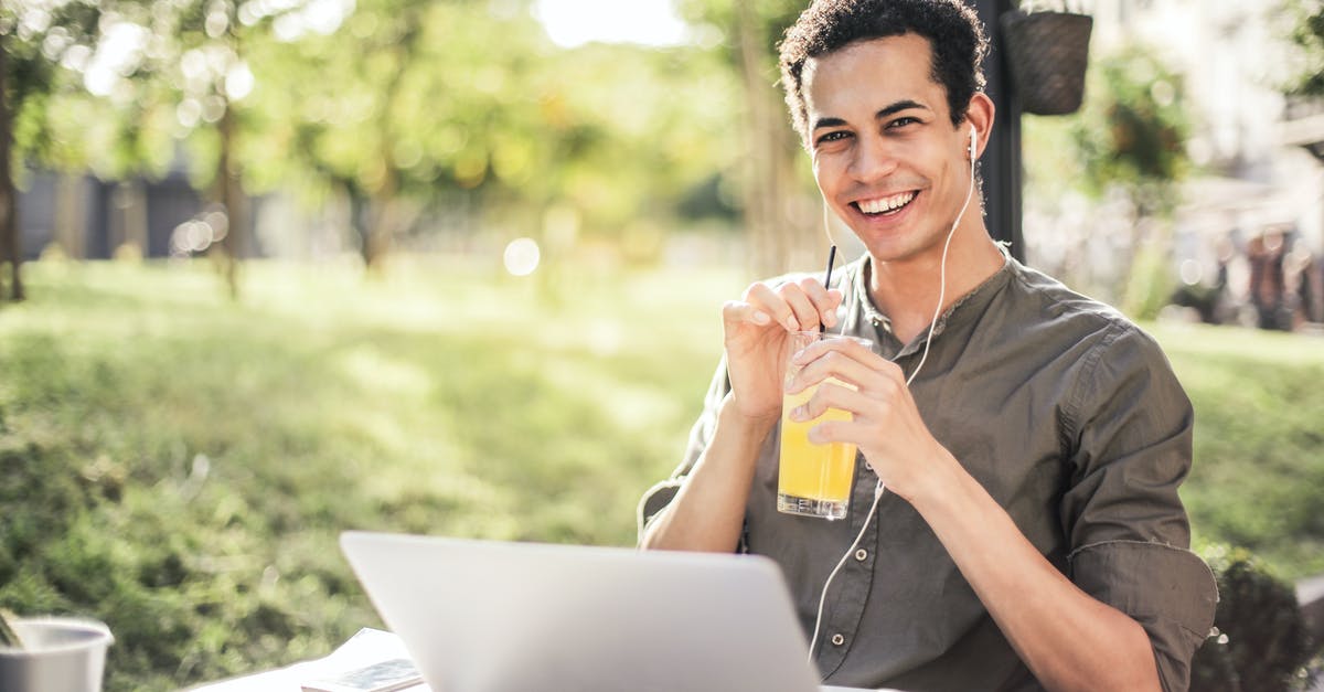 How can I chill ice coffee quickly? - Cheerful guy with laptop and earphones sitting in park while drinking juice and smiling at camera