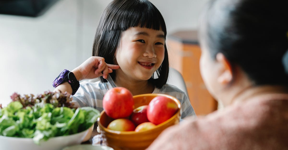How can I create this coconut milk, fruit, and veggie snack at home? - Delighted cute ethnic little girl smiling while sitting at table and eating delicious fresh vegetables and fruits with unrecognizable mother