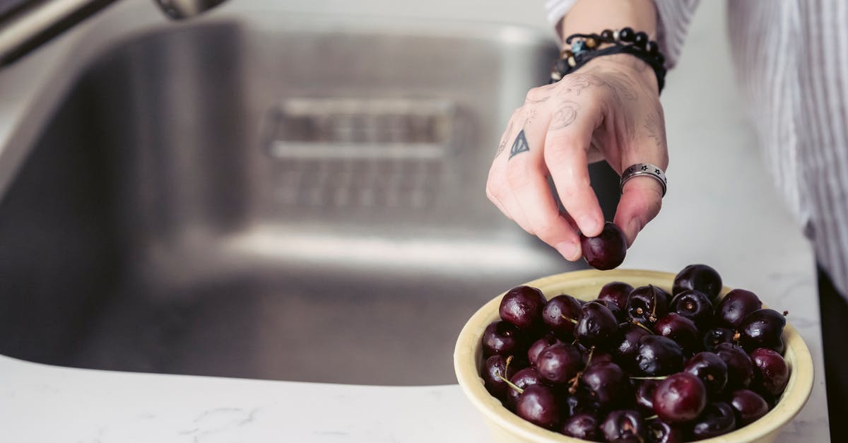 How do I keep fruit from sinking to the bottom of my cake? - From above of crop unrecognizable tattooed person in casual clothes standing near sink in kitchen and eating bowl of fresh washed cherries