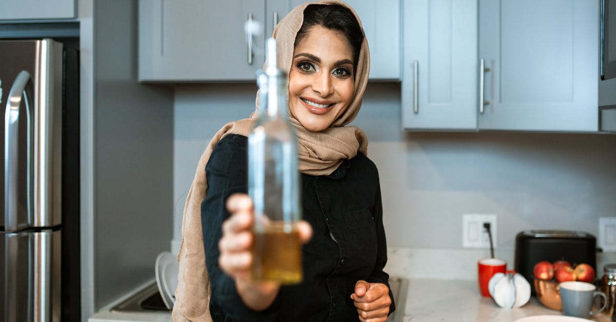 How do I prevent mold growth in homemade chili-infused olive oil? - Happy ethnic female in headscarf showing bottle of organic olive oil and looking at camera with smile while cooking in contemporary kitchen