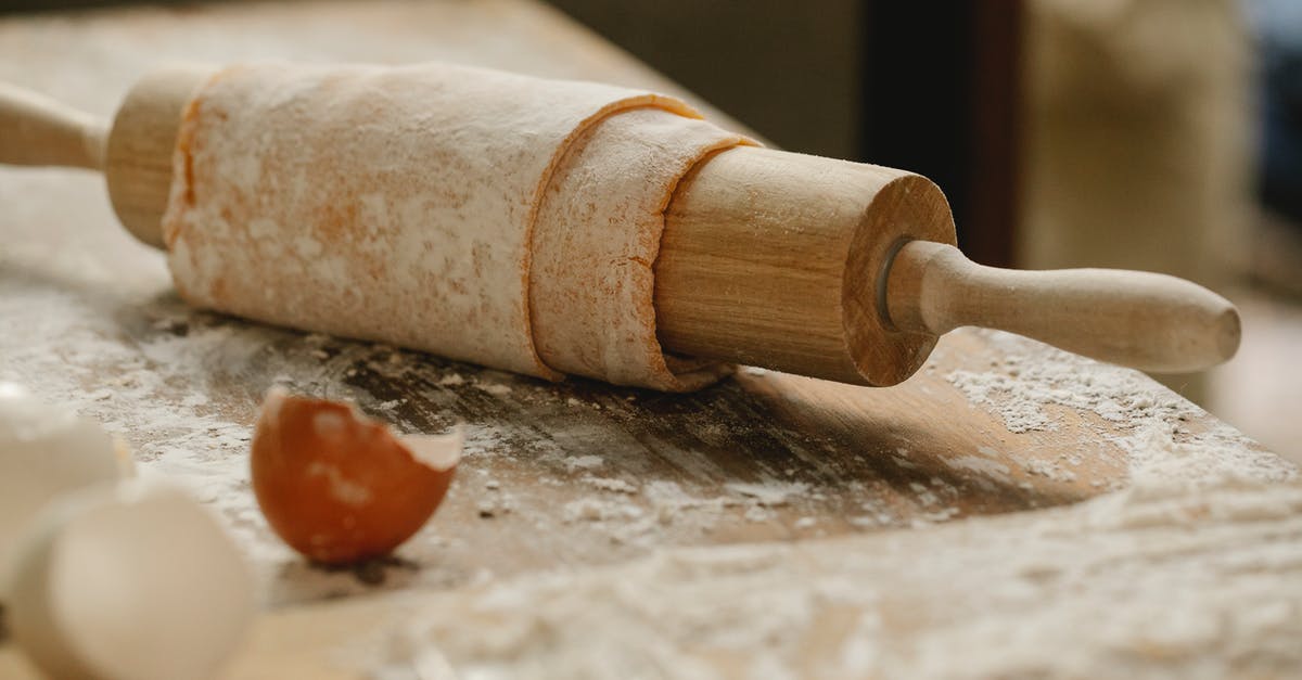 How do I roll or wrap up pasta into a log-like shape for presentation? - Rolling pin wrapped with uncooked sheet of dough placed on wooden table sprinkled with flour near eggshells in kitchen on blurred background