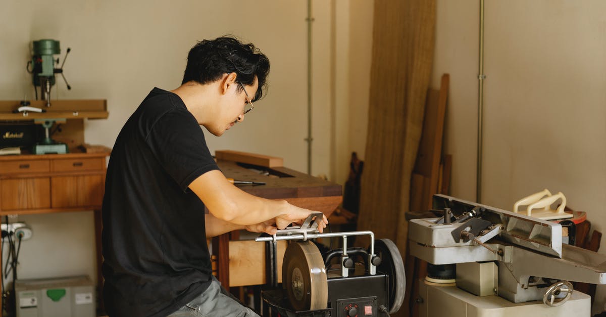 how do I sharpen grinder blade - Side view of young concentrated Asian male joiner working on grinder with sharp wheels while sitting on stool in workshop