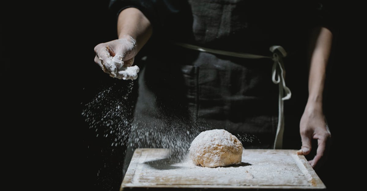 How do professional chefs keep food thermometers clean between uses? - Unrecognizable baker in uniform standing at table and sprinkling flour in dough while cooking against black background