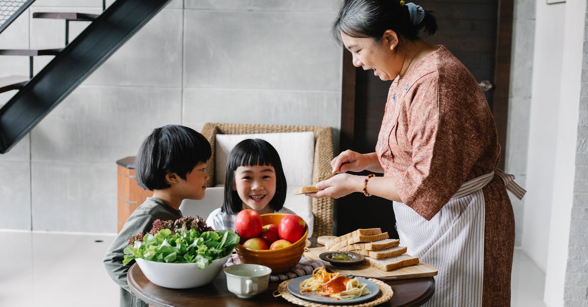 How do you make Welch's fruit snacks? - Side view of positive senior Asian female in apron spreading butter on bread while preparing breakfast for funny little grandchildren sitting at round table in cozy kitchen