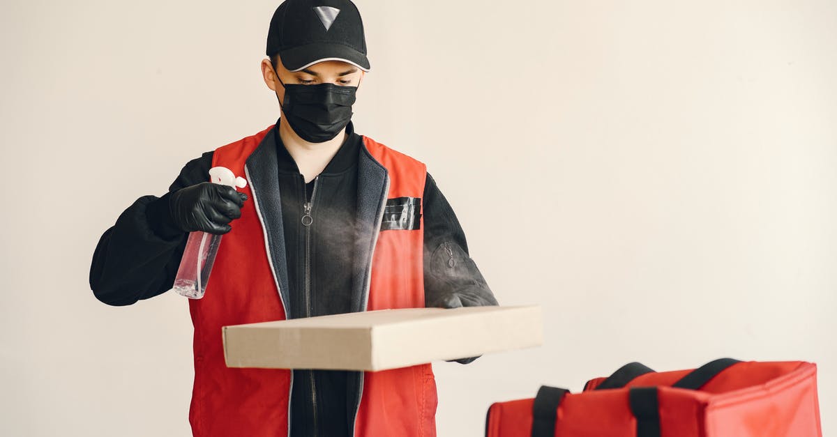 How do you protect a pizza peel? - Male courier in cap and uniform wearing medical facial mask and gloves spraying antiseptic on carton pizza box against white background near thermo bag illustrating safety food delivery concept