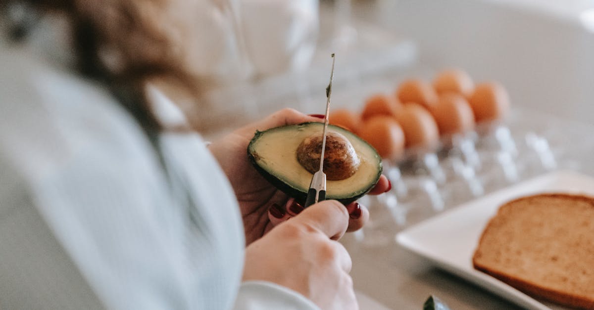 How do you remove the pit from a mango? - Crop anonymous female cook with knife preparing avocado while standing at table with eggs and toasts in light kitchen at home