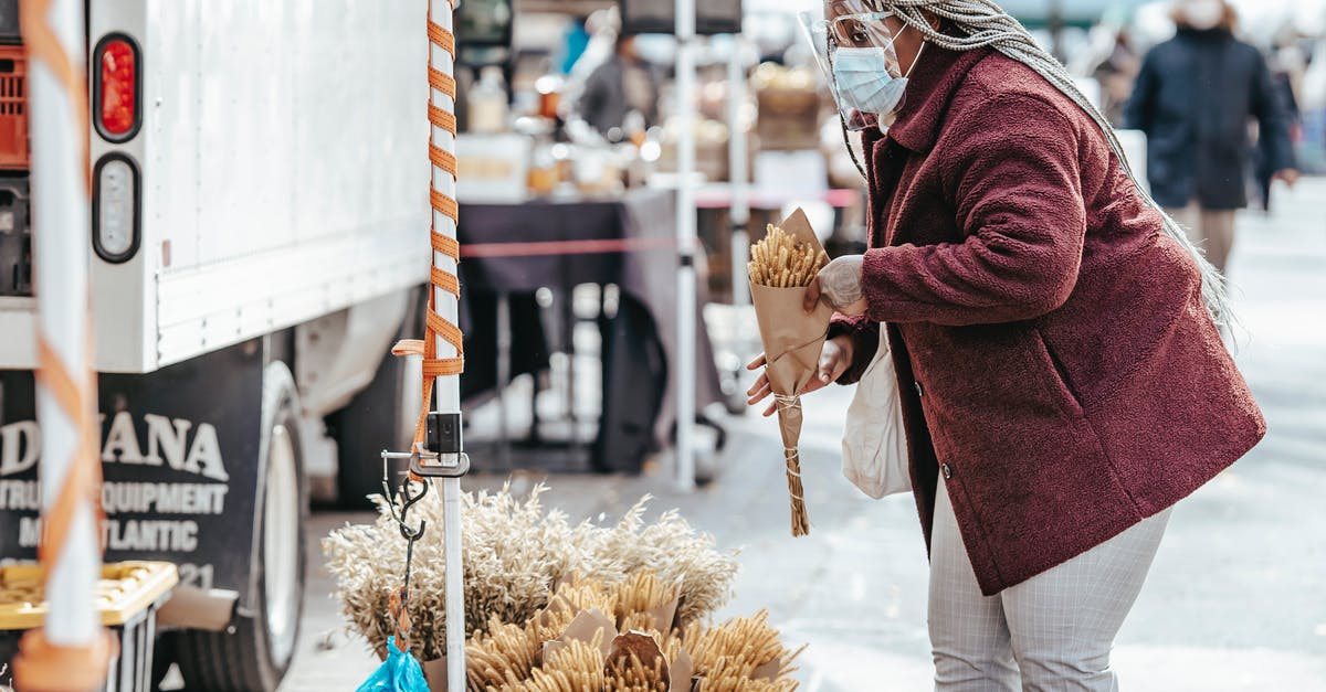 How should I choose corn? - Black adult lady in warm coat and protective mask standing in street market and choosing bouquet of dried corn in daytime
