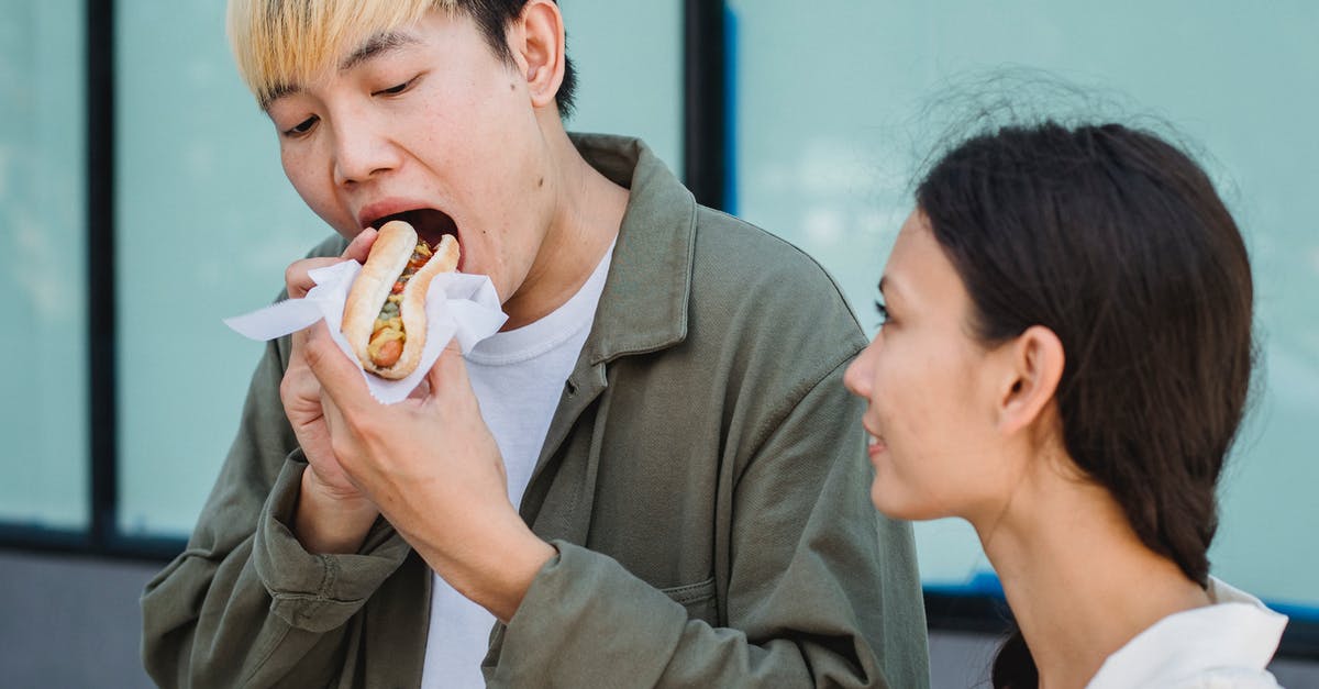 How to calculate the calorie content of cooked food? - Content Asian male in casual outfit eating delicious hot dog while standing on sunny street near attractive girlfriend
