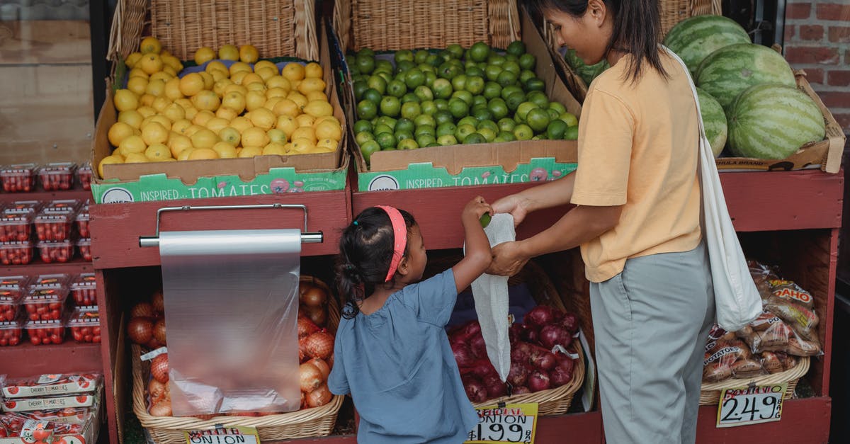How to pick a good mango at a local store? - Back view of Asian girl picking ripe limes from box to eco friendly bag while shopping with mother in market
