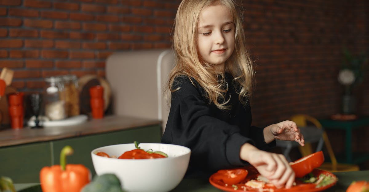 How to remove unwanted aftertaste from food - Cheerful little blond child preparing salad while removing pepper seeds and standing near table with fresh vegetables behind brick wall in apartment