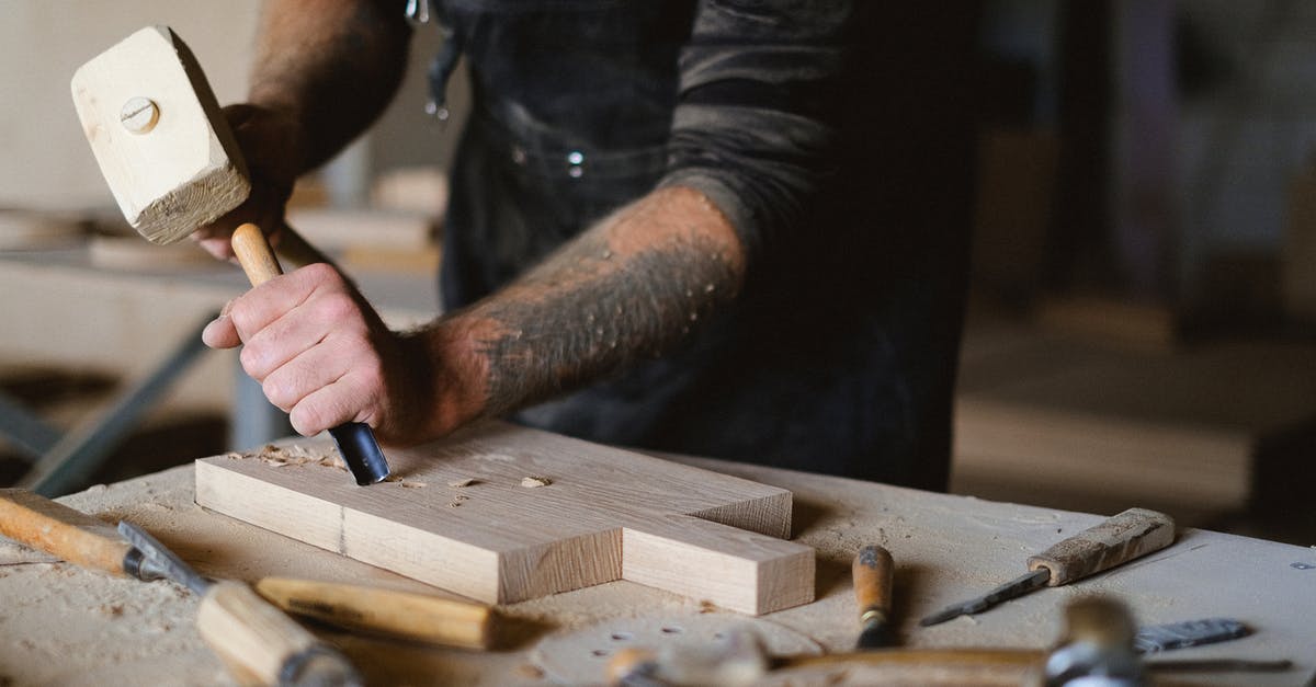 How to shell macadamias without a special tool? - Unrecognizable male carpenter using forged chisel and wooden hammer while creating pattern on lumber board at table in workshop on blurred background