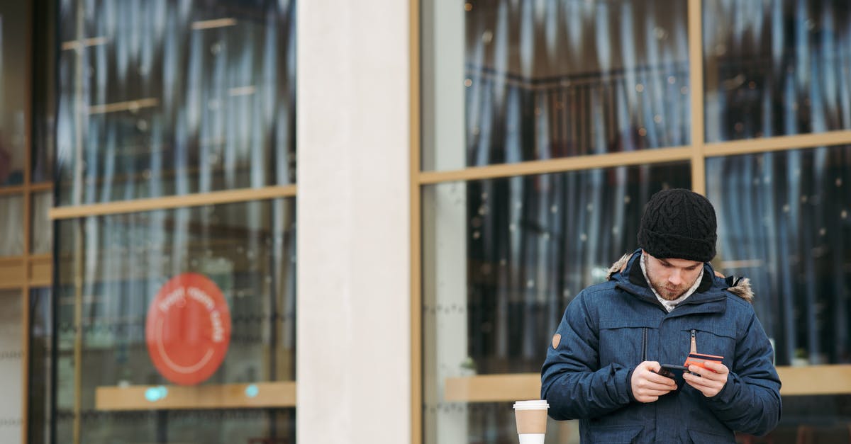 How warm must dough be in order to rise? - Focused young male in warm outerwear and hat standing near modern building with takeaway coffee and using smartphone while making online shopping with credit card