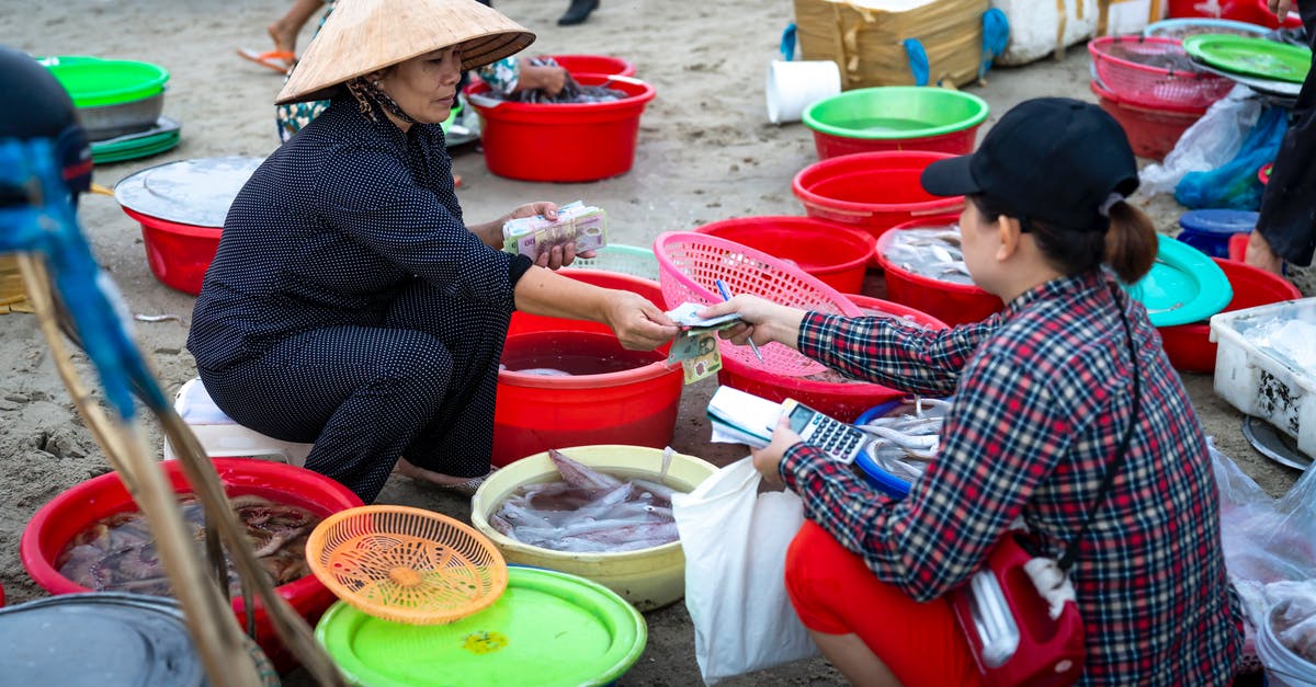 In England, can I buy fresh fish directly off the boat? - Asian female in traditional authentic Vietnamese hat sitting on stool in street fish market and selling seafood to female customer