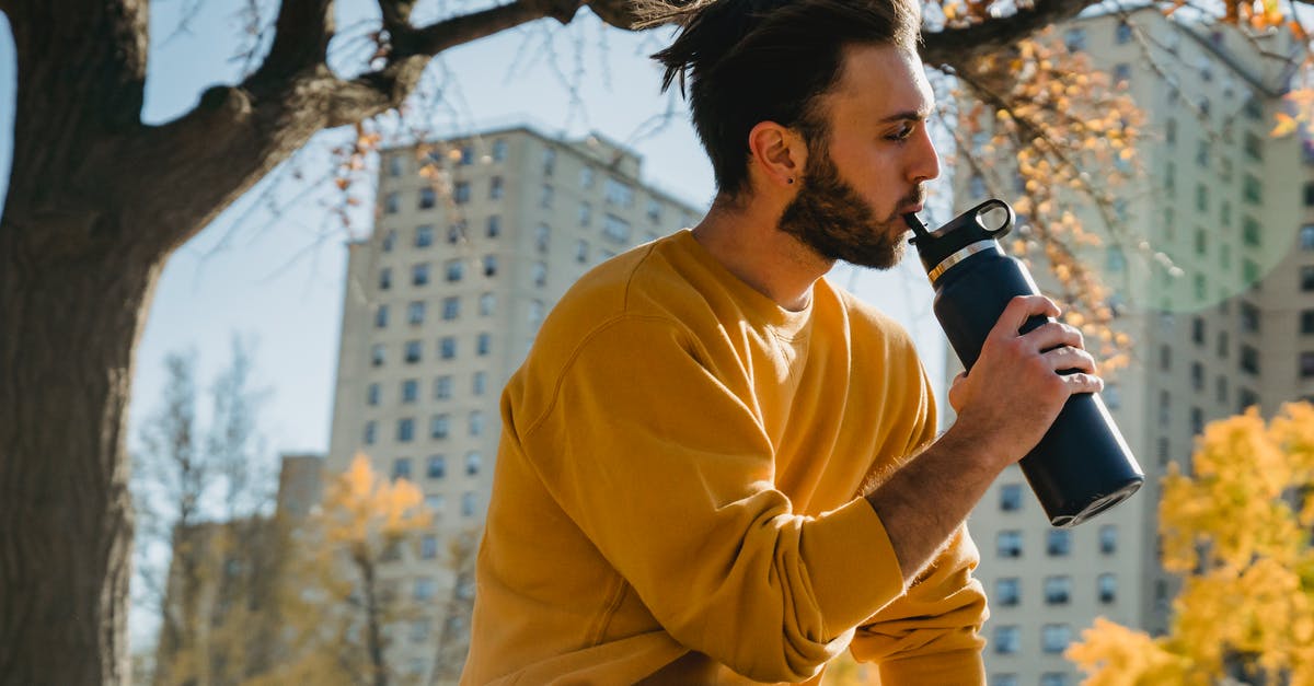 Is it ok to keep reusing a water bottle indefinitely? - Side view young calm male in orange sweater drinking water from reusable bottle and looking away while sitting in autumn city park