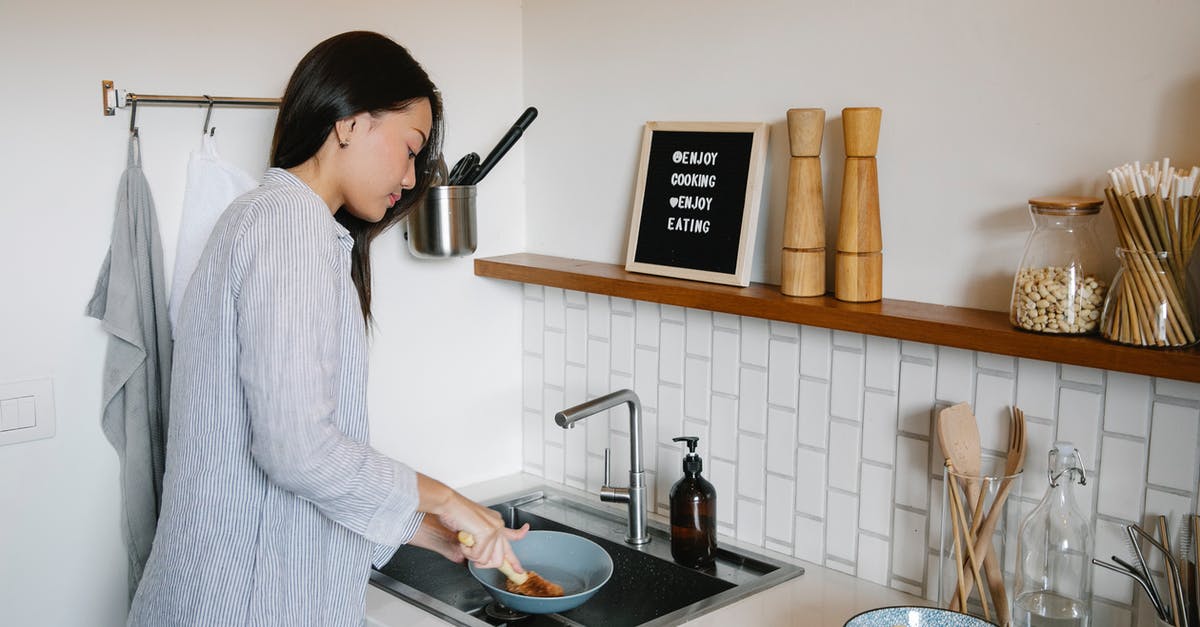 Is it okay to wash your hands in the kitchen sink washwater (with the dishes)? - Side view of Asian female with brush washing dishes at sink while doing housework in light kitchen with supplies at home