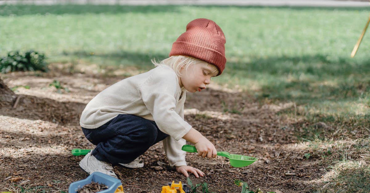 Is there a reason to have a plastic bowl? - Side view of little boy in casual clothes and brown hat playing with plastic toys in backyard