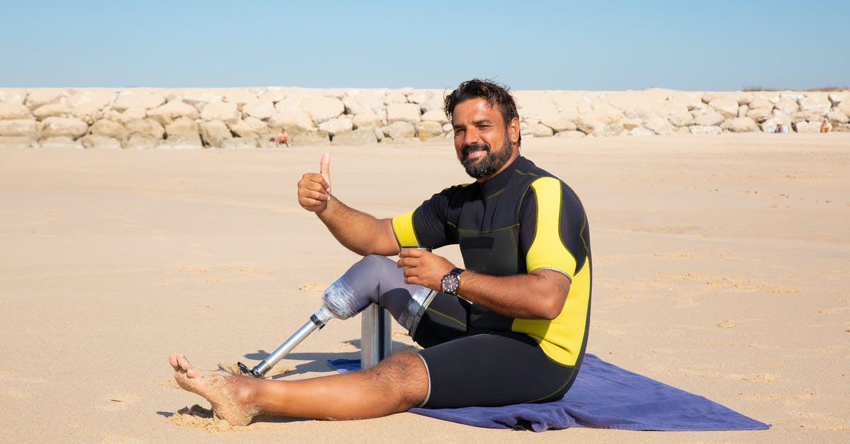 Is there some drink like panaché available in the Netherlands? - Side view full length handsome ethnic man with leg prosthesis resting on sandy seashore and showing thumb up gesture while looking at camera with smile