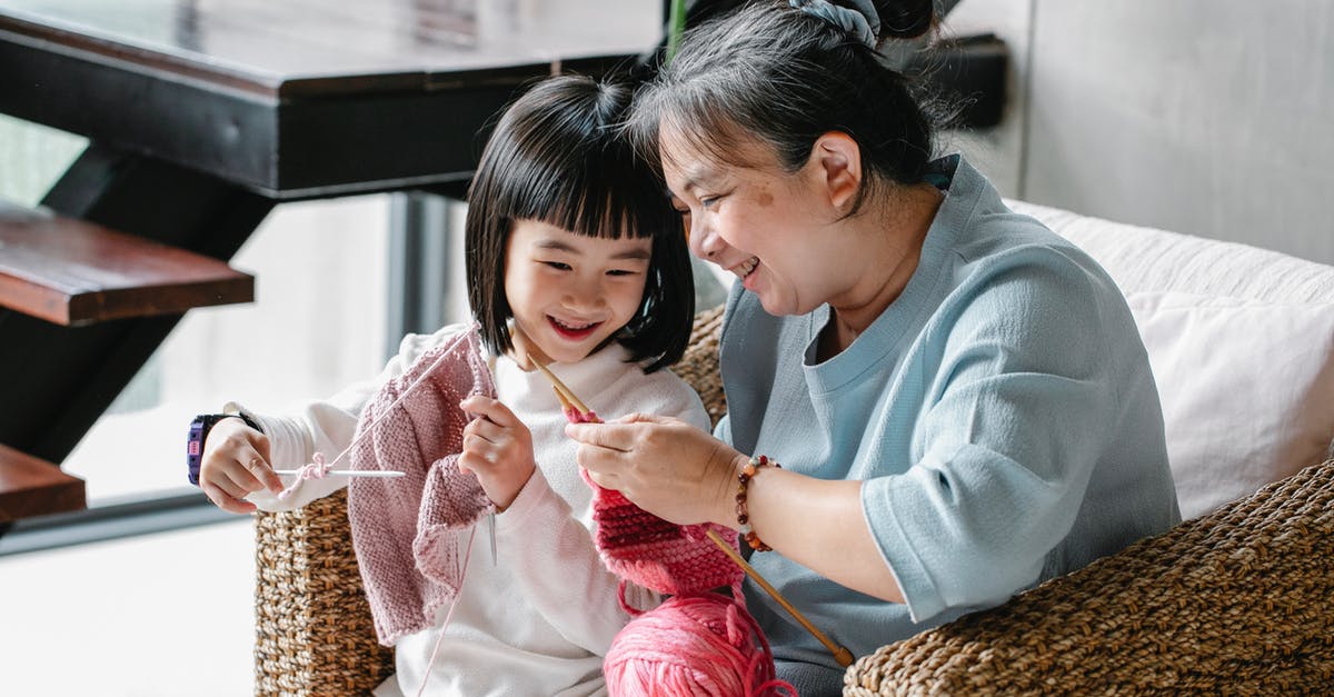 Is this some kind of warming oven? - Cheerful senior Asian woman sitting in armchair and helping curious little granddaughter  to knit warm clothes with yarn in daylight