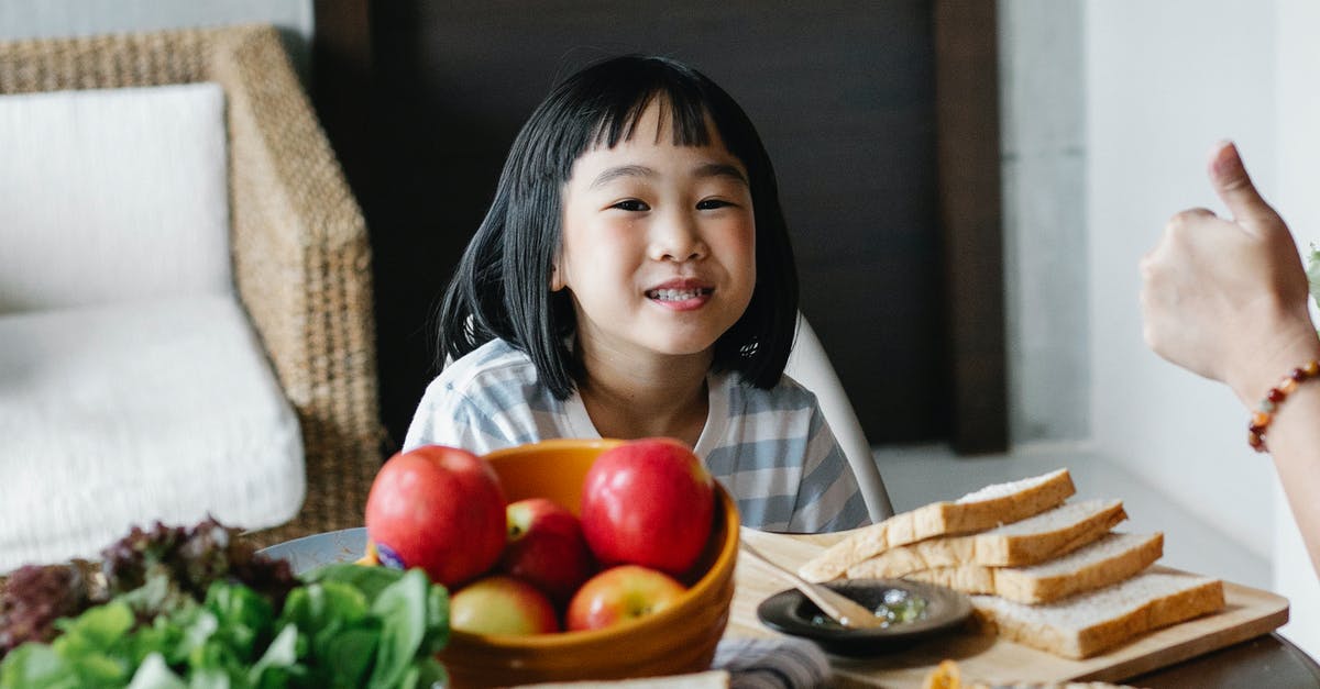 Kind of apples for apple purée / apple sauce - Positive cute little Asian girl sitting at table with bowl of apples and green salad served with sliced bread and spaghetti during lunch at home