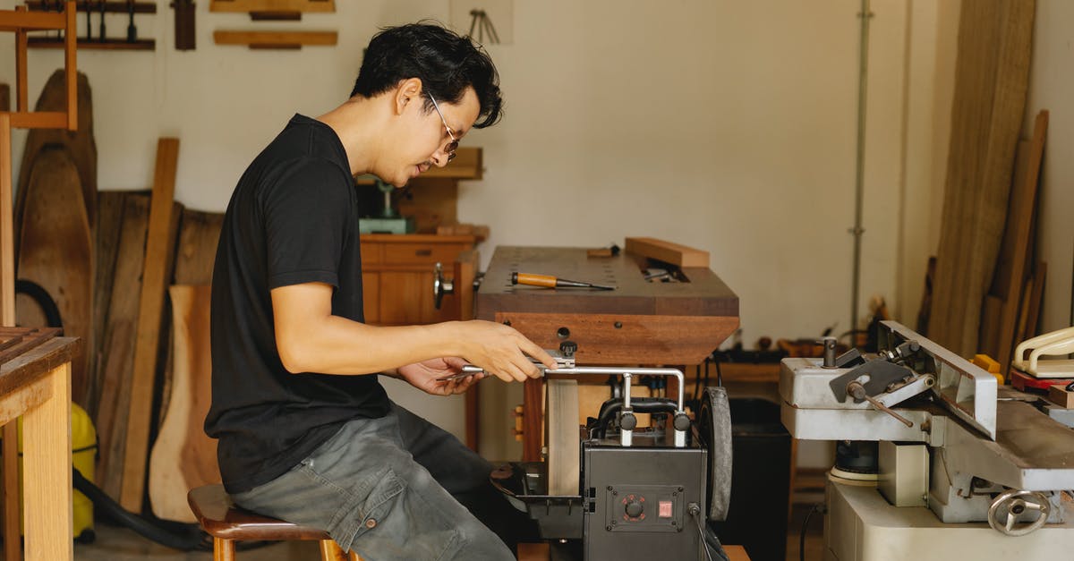 Knife sharpening equipment - Side view of concentrated skilled ethnic male master sitting at electrical sharpening machine with sharp knife during work in joinery