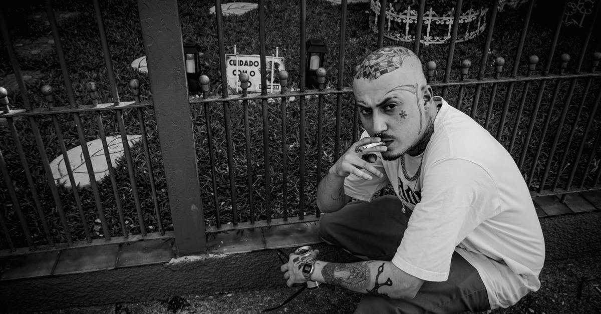 Looks curdled, but it wasn't - my bad alfredo - Black and white from above of serious tattooed male smoking cigarette while sitting on haunches near metal fence on street