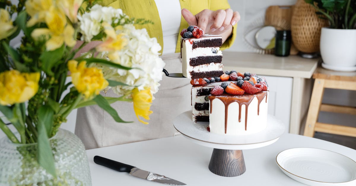 Making cake balls with a not-so-sweet frosting - Crop unrecognizable female confectioner in casual clothes and apron serving yummy homemade cake with white cream and assorted fresh berries standing in light kitchen decorated with flowers vase and plant