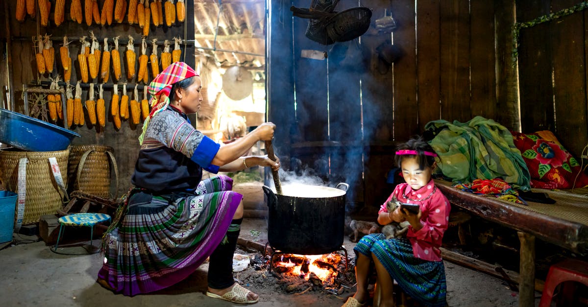 Making Tamales using a corn meal alternative - Side view of Asian woman sitting and preparing food in cauldron against wall with corn while little daughter surfing smartphone in old wooden barn