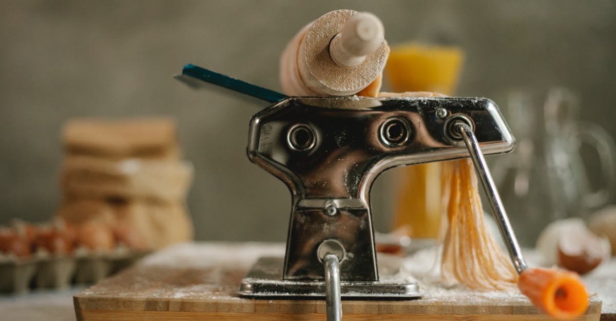 Manual noodle press pasta maker sticking - Stainless modern pasta maker instrument with handle and cut dough placed on table with products on blurred background  in kitchen