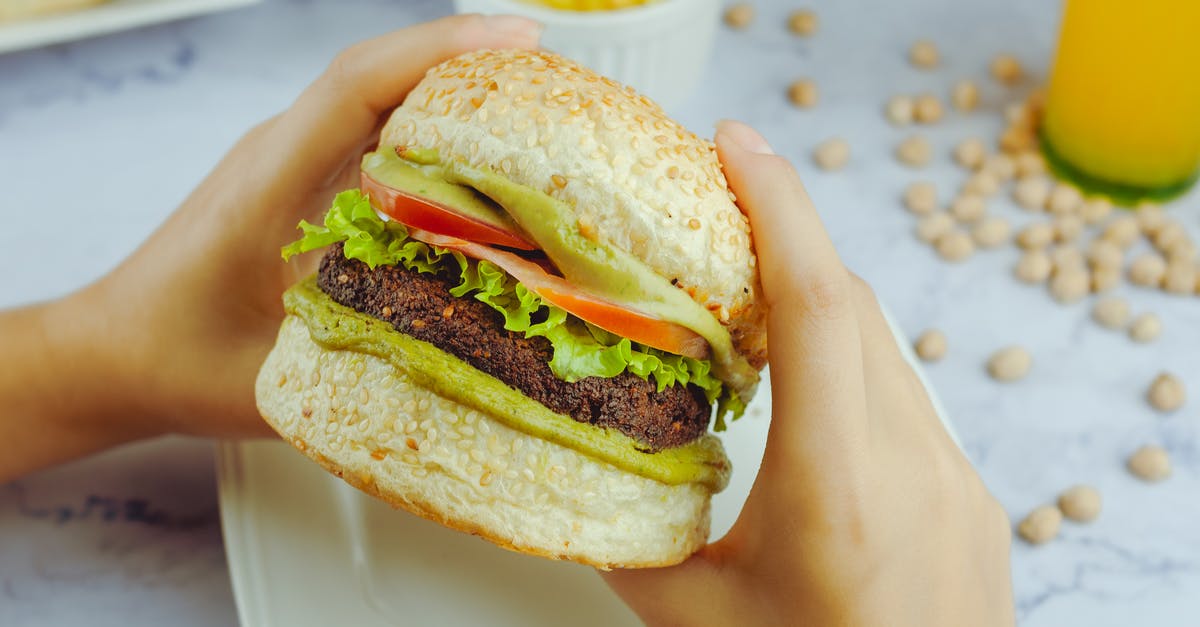 Meat juice reabsorption sous vide - Crop person eating big appetizing hamburger with fried cutlet between fresh vegetables and green sauce at table with juice and French potatoes