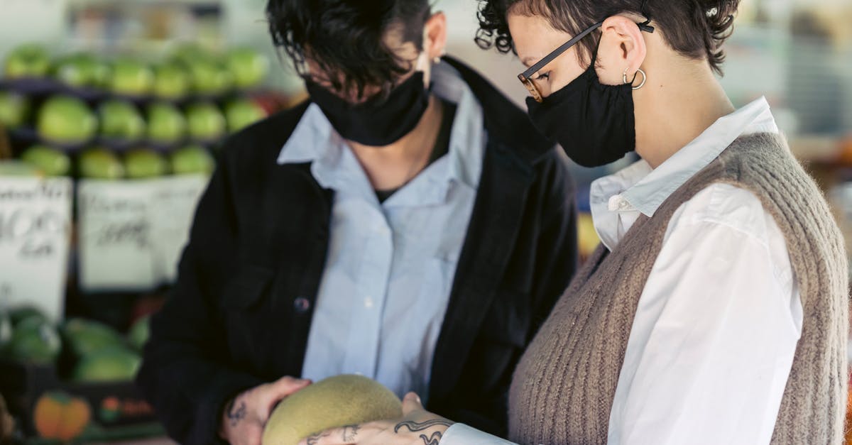 Melon buying tips - Crop unrecognizable female friends with short dark hair in casual clothes and protective masks examining melon while purchasing products in grocery market