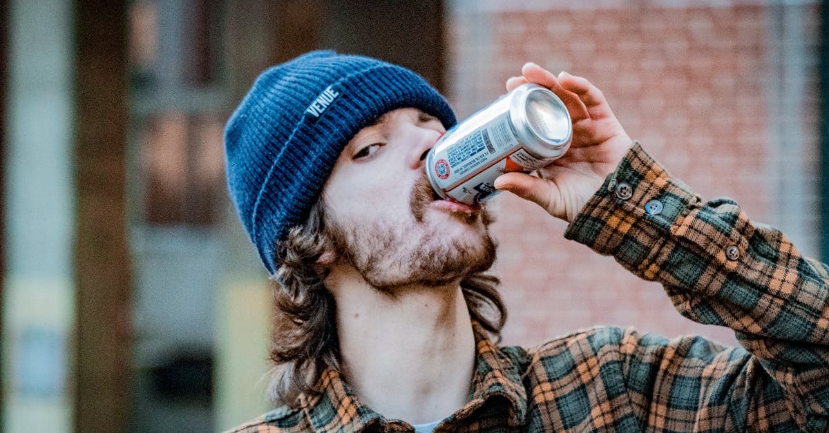 Mixed drink with tonic water already added? - Calm young male in casual checkered shirt and hat enjoying cold drink from can while standing on street and looking at camera
