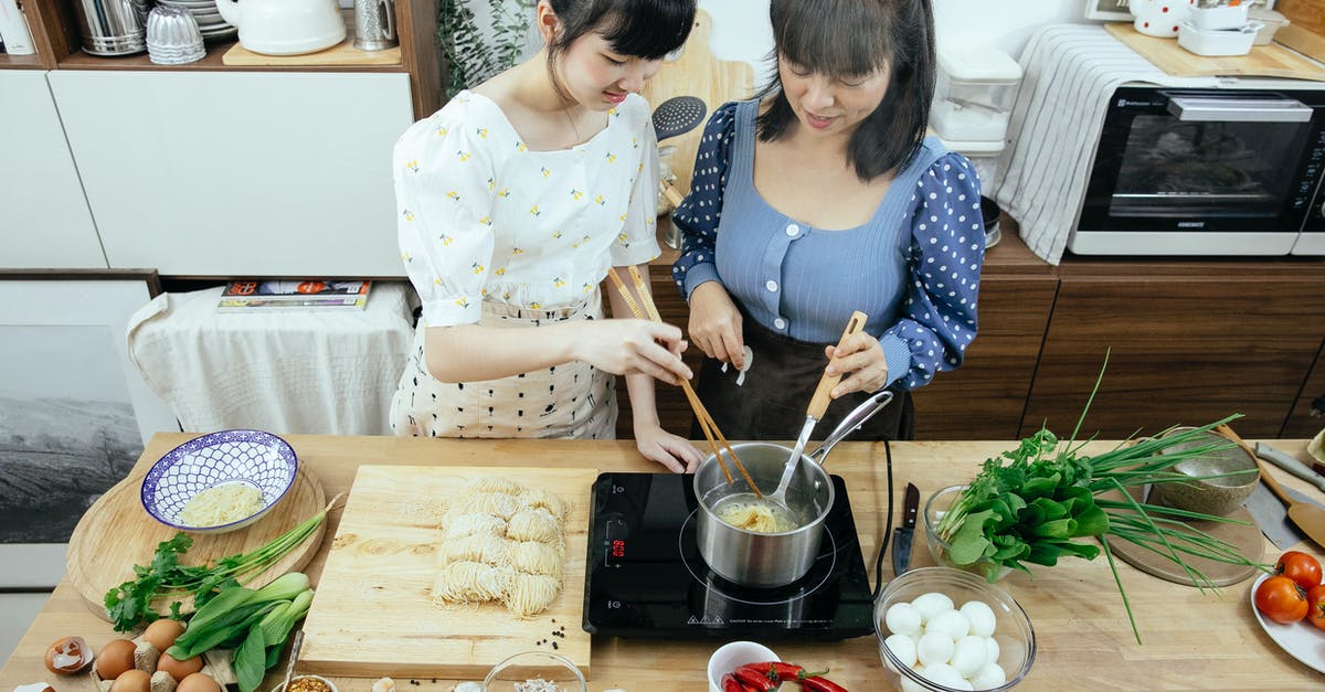 Mysterious Disintegrating Udon noodles - High angle glad Asian females in stylish aprons with chopsticks boiling homemade noodles in saucepan on tabletop stove while cooking Asian dish together in contemporary kitchen