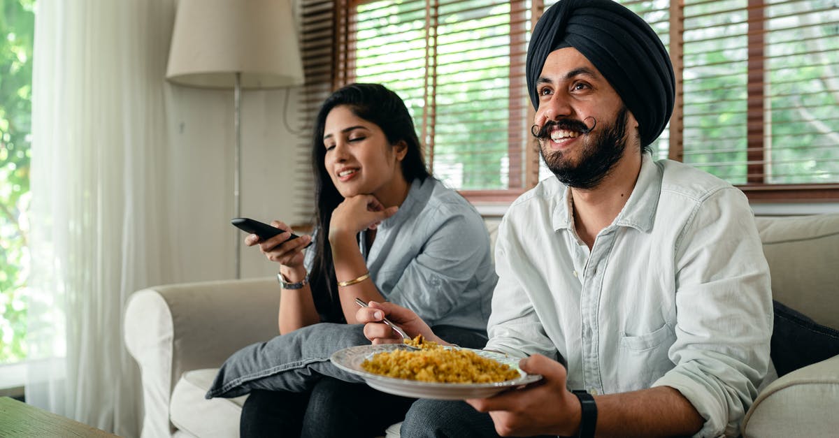 Name of the snack (possibly Indian) - Positive young bearded man in casual wear and turban eating traditional saffron rice on sofa while wife switching channels on TV with remote controller