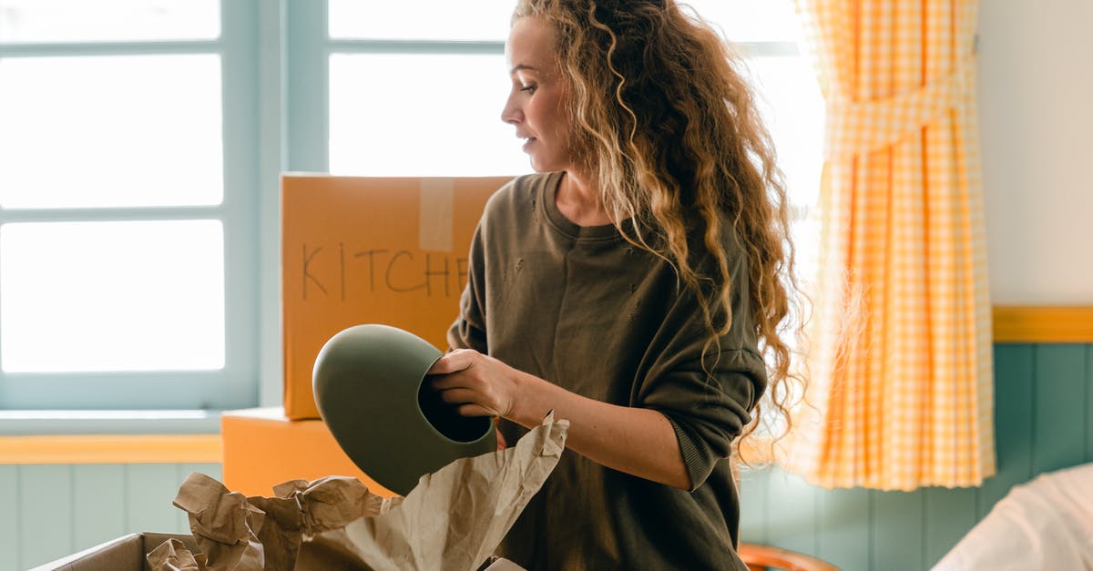 Navigating the different types of cookware - new kitchen advice - Dreamy female in casual wear taking ceramic vase out of parchment while standing near cardboard box with KITCHEN inscription in back lit preparing to move into new house