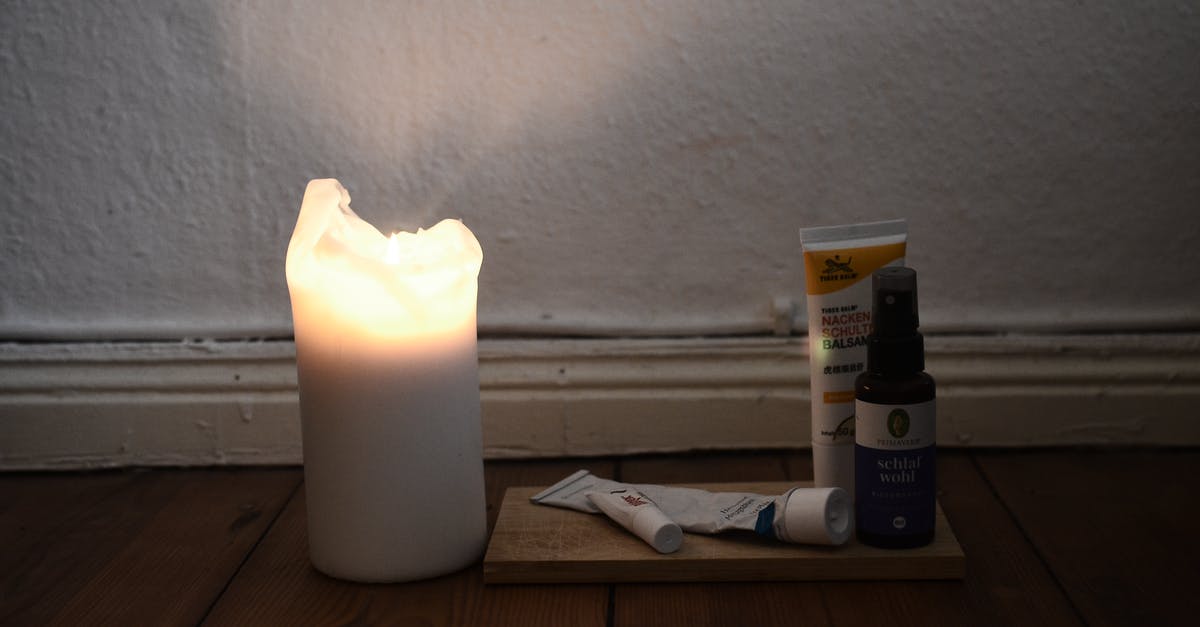 New oil vs old oil - White burning candle placed on wooden floor with vials and tubes for skincare daily routine against white shabby wall in room at low light