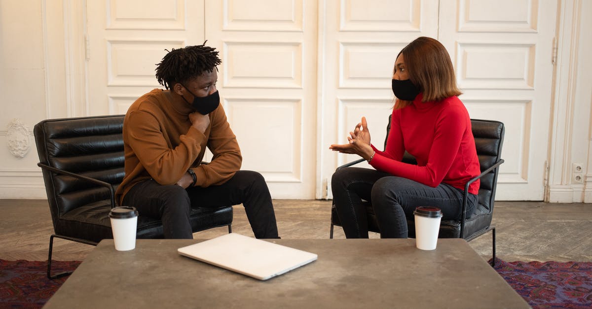 Normal coffee vs decaf - Focused African American colleagues in protective masks looking at each other while having conversation about work at table with laptop and paper cups of takeaway coffee