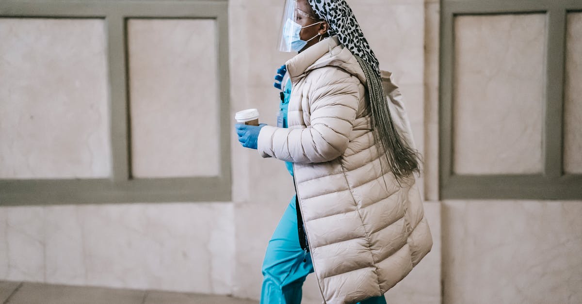 Normal coffee vs decaf - Side view calm adult African American nurse in uniform and warm clothes wearing medical mask face shield and latex gloves walking with coffee to go on street