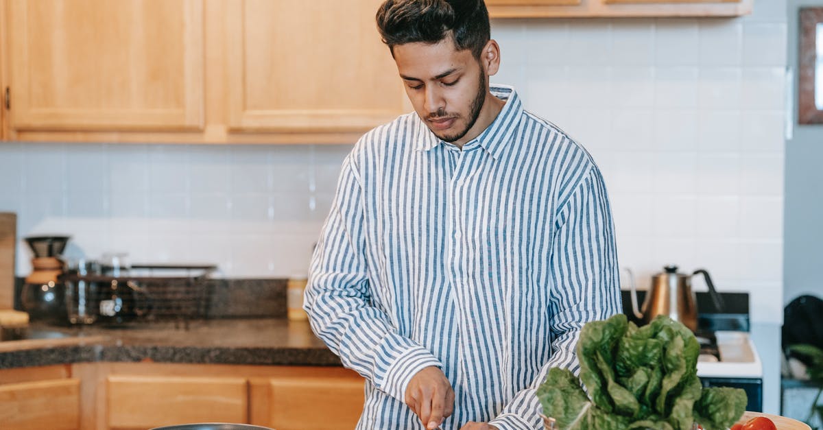 Nutrient impact of squeezing water out of frozen chopped spinach - Bearded ethnic male in striped shirt cutting fresh tomato with knife against spinach leaves on table at home