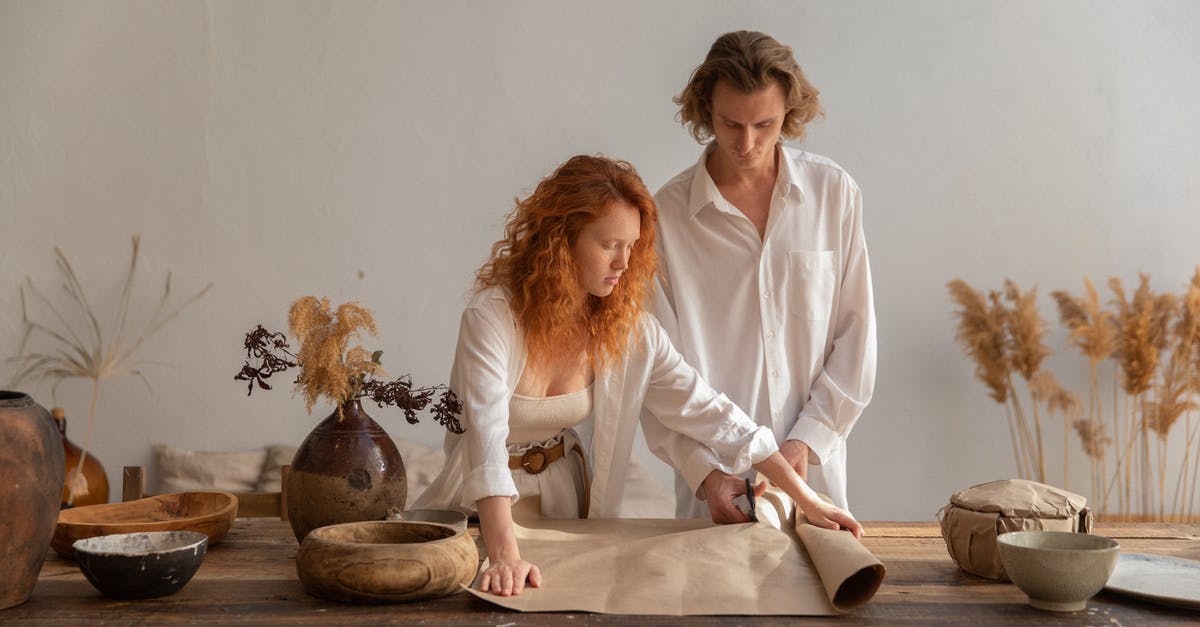 On preserving dried ginger - Concentrated man and redhead woman standing near wooden table with craft paper placed near bowl during packaging in cozy workshop