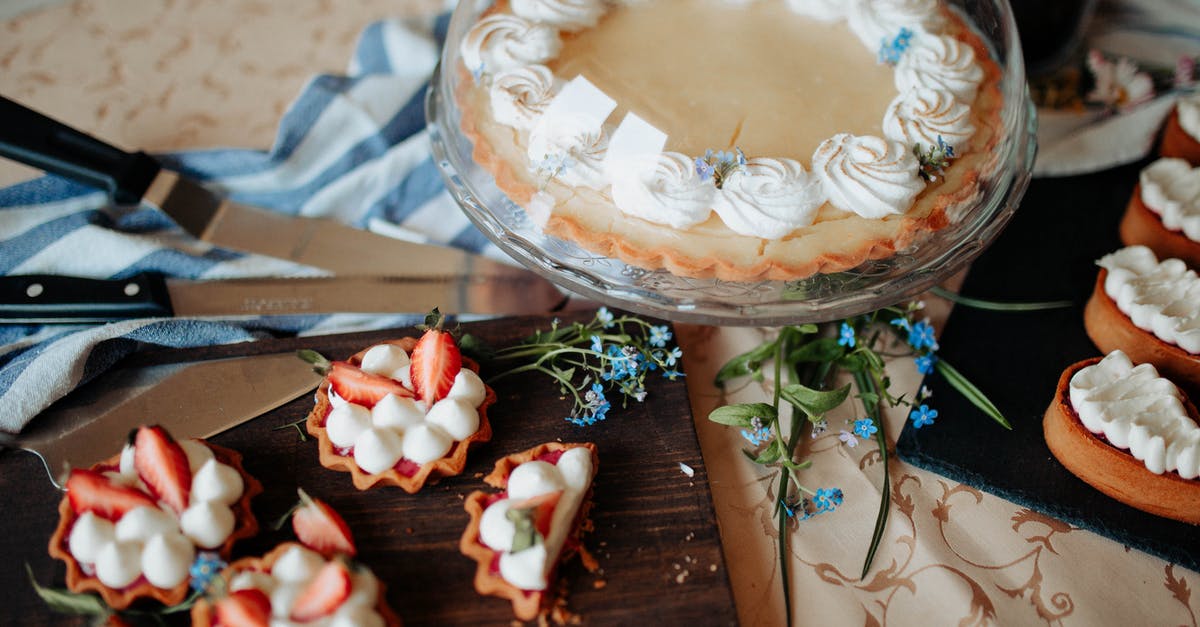 Pie with 2 different fillings - Pie decorated with cream on tray with glass cap near flowers and different desserts on cutting boards near knives on table in bright kitchen