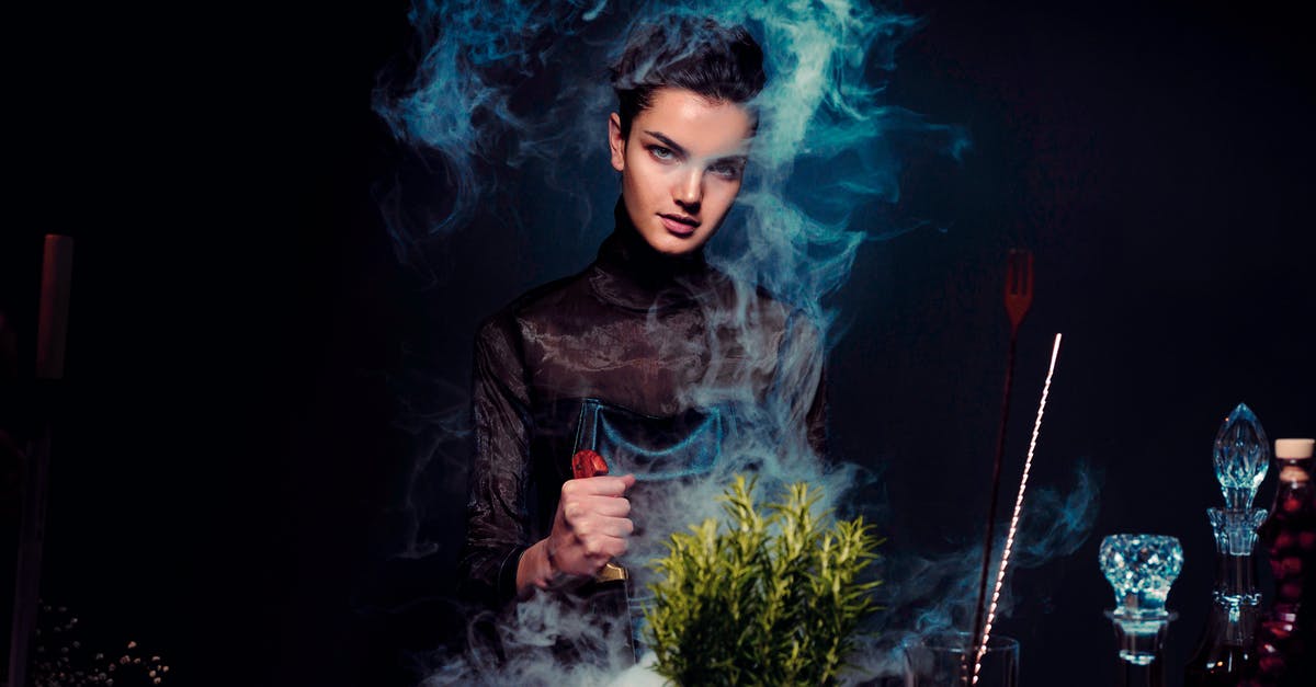 Preparing Salsa with a Magic Bullet - Graceful young female alchemist with knife in hand in black outfit preparing potion from various herbs among smoke in dark room