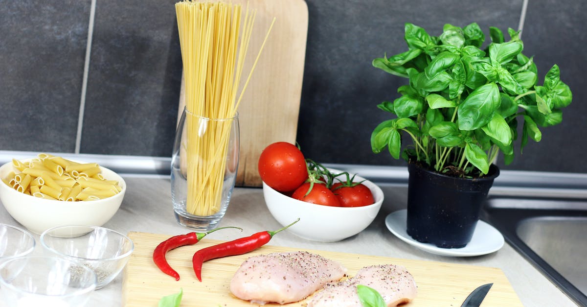 Preparing slow cooked meats for the next day - Raw Chicken Breast Seasoned With Peppers Beside Red Chili, Basil, Bowl of Tomatoes, and Raw Pastas on Table