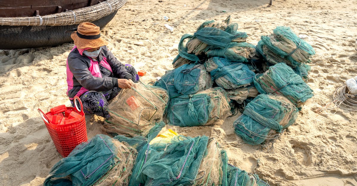 Prevent gummi bears from drying out - From above of anonymous ethnic female in hat and mask tying fishing net against oriental boats on beach in sunlight