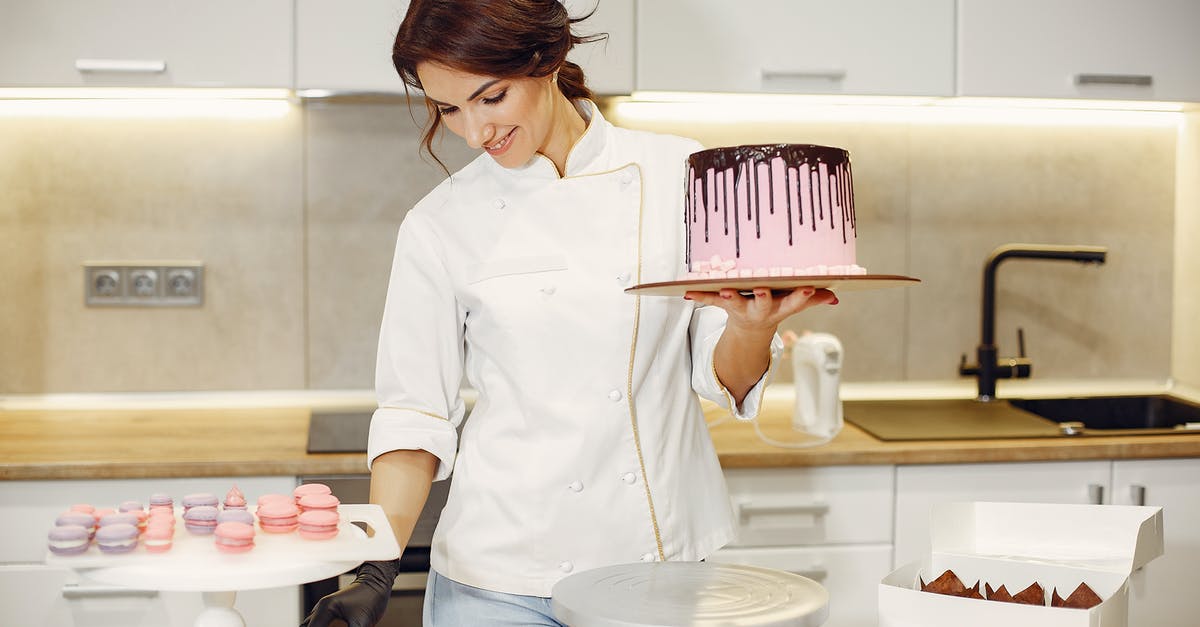 Putting a solid chocolate bar inside cake dough: what's the outcome? - Positive female confectioner in uniform holding pink cake on cardboard with chocolate drips while decorating with colorful marshmallows standing in kitchen