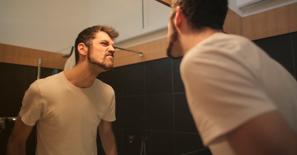 Question about mirror glazes and freezing - Low angle side view of young bearded male in casual shirt standing in bathroom and looking at with frown mirror in morning