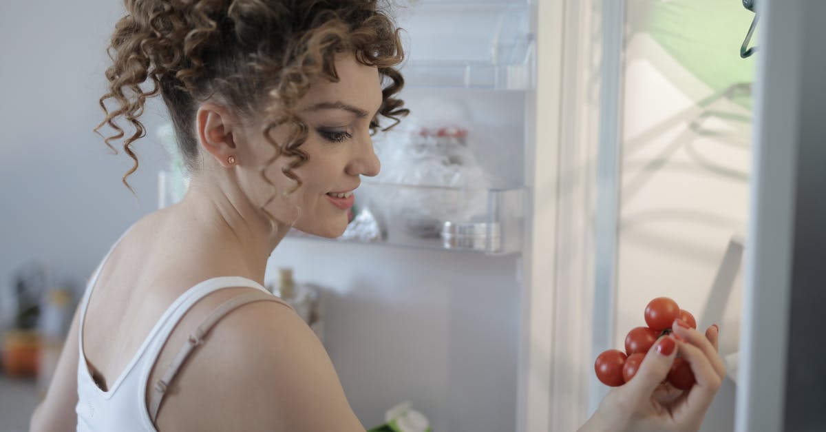 Refrigerator Fruit/Vegetable Bins - Side view of content female in casual wear standing in kitchen near opened fridge and picking fruit for breakfast