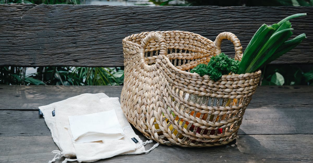 Reuse of Food Grade Tins - Wicker basket with heaped organic vegetables placed on wooden bench near reusable grocery bags in lush garden