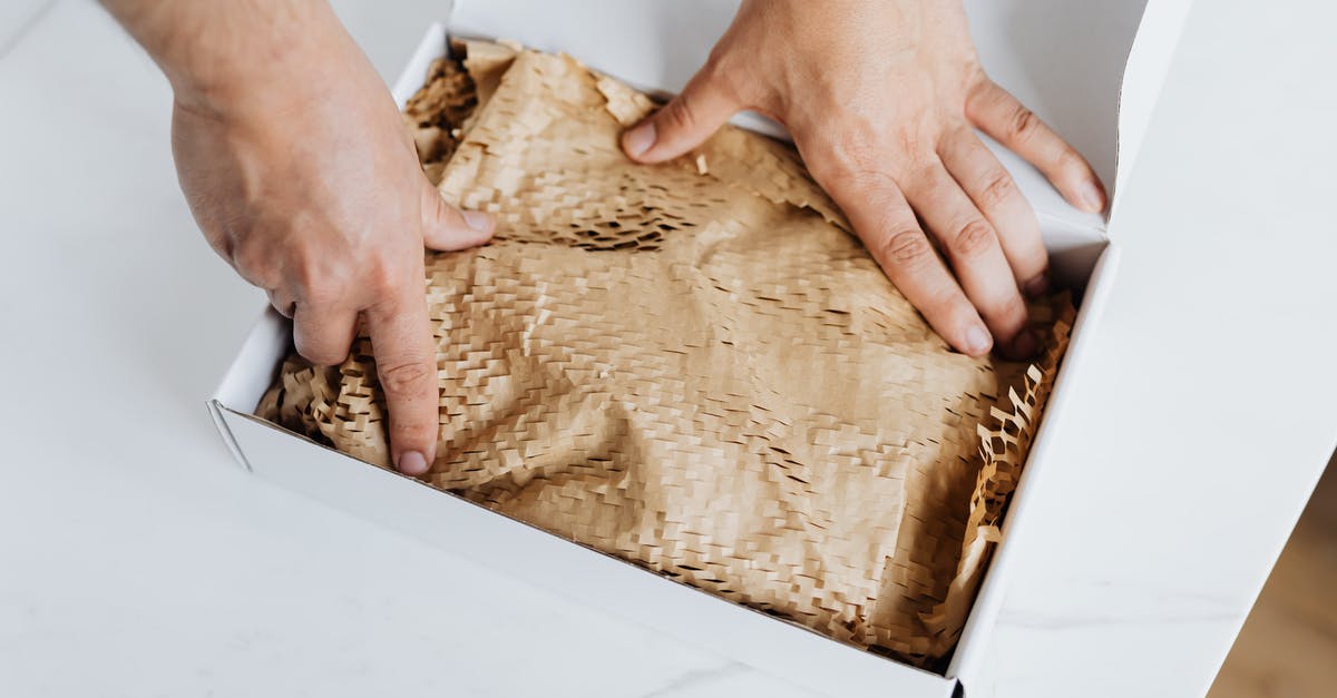 Safety of producing home malted flour - From above crop adult male packing ordered product or gift in white cardboard box with craft paper for safety postal delivery at white marble table