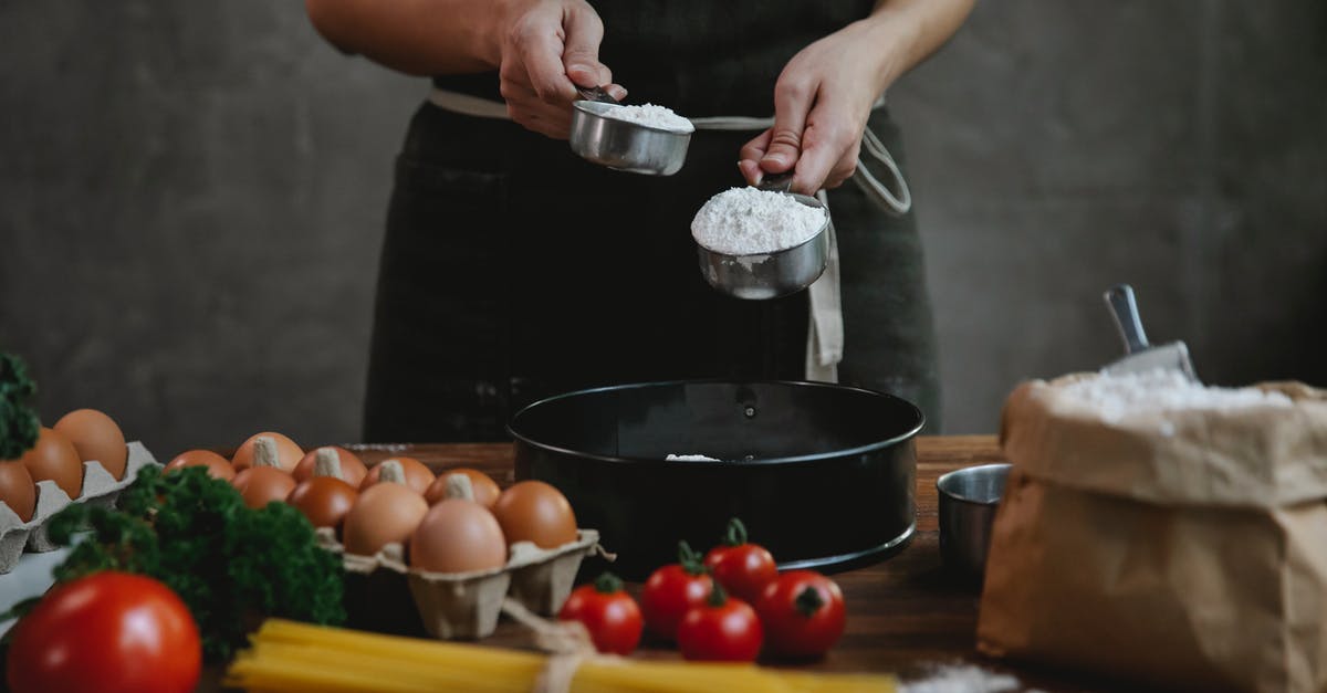 Sanitising Black chef "whites" - Crop faceless person in black apron standing near counter and adding flour in bowl while making homemade pasta with eggs and spaghetti near tomatoes and herbs